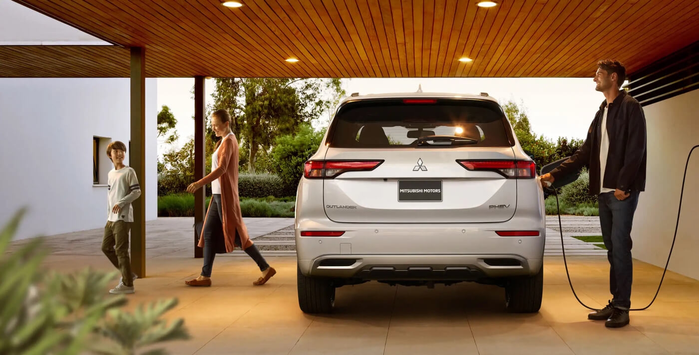 Un homme qui branche son Mitsubishi Outlander PHEV 2024 pendant que sa famille rentre dans la maison.